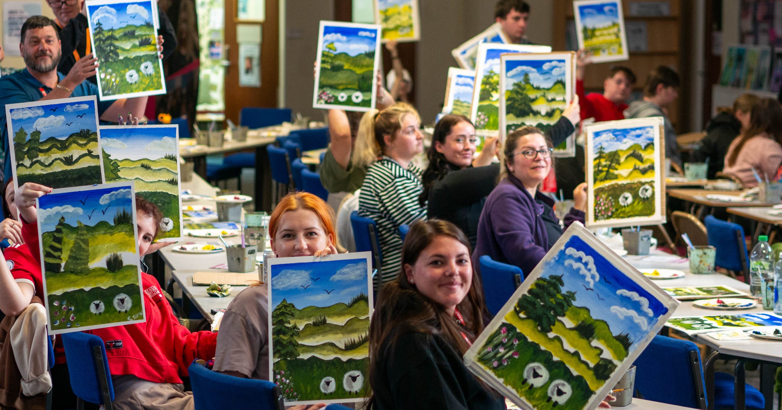 Students and staff holding up finished paintings on canvas of Welsh hill scene with sheep.