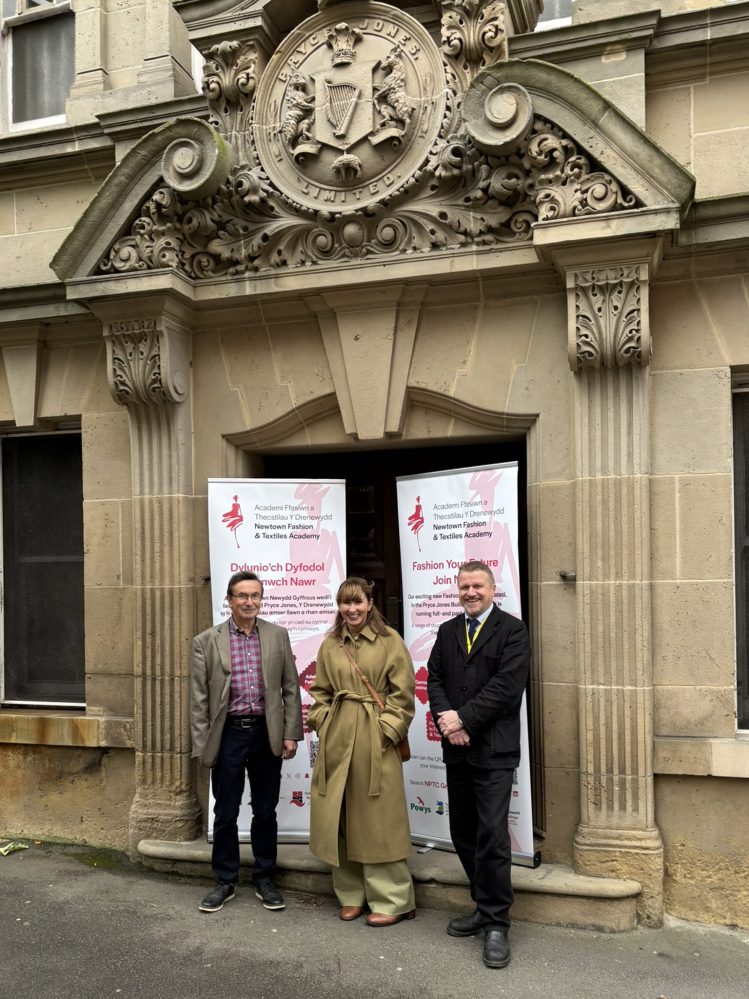 Vice Principal Gemma Charnock, Campus Manager Steve Cass and Councillor David Selby standing outside the entrance to the Pryce Jones Building with two pull-up banners with Newtown Fashion & Textiles Academy branding.