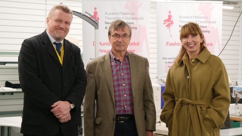 Vice Principal Gemma Charnock, Campus Manager Steve Cass and Councillor David Selby standing in a workshop in the Pryce Jones Building with two pull-up banners with Newtown Fashion & Textiles Academy branding.