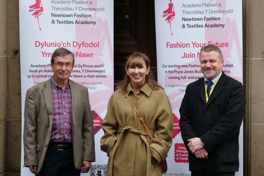 Vice Principal Gemma Charnock, Campus Manager Steve Cass and Councillor David Selby standing outside the entrance to the Pryce Jones Building with two pull-up banners with Newtown Fashion & Textiles Academy branding.